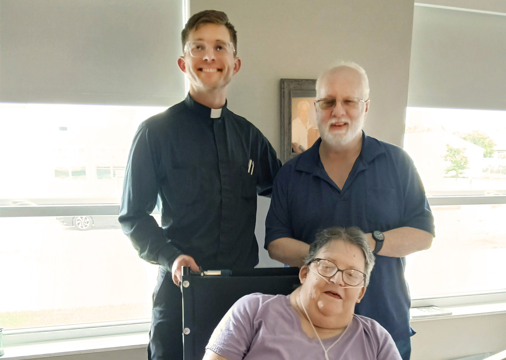 Three people pose indoors by bright windows: two standing behind a seated person in a wheelchair, all wearing glasses. 