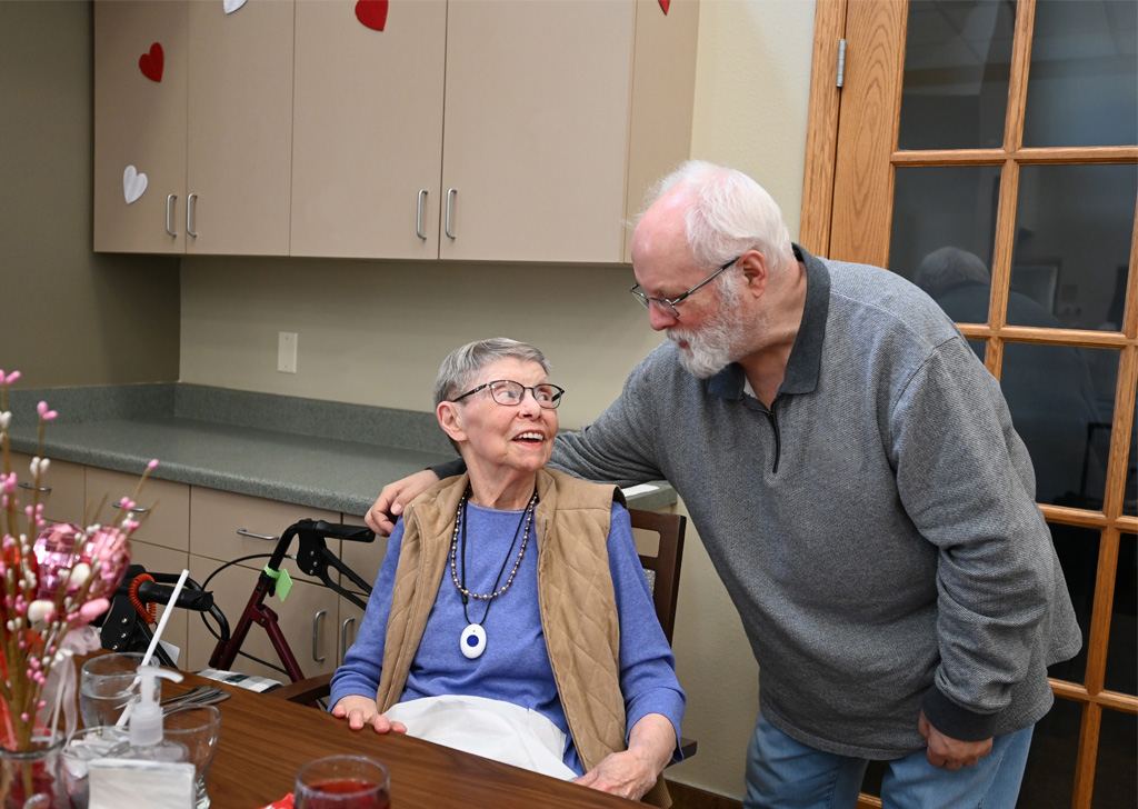 An older adult sits at a table in a dining area while another person stands beside and leans in with an arm around the seated person; a walker, cabinets, and a glass door are visible in the background.