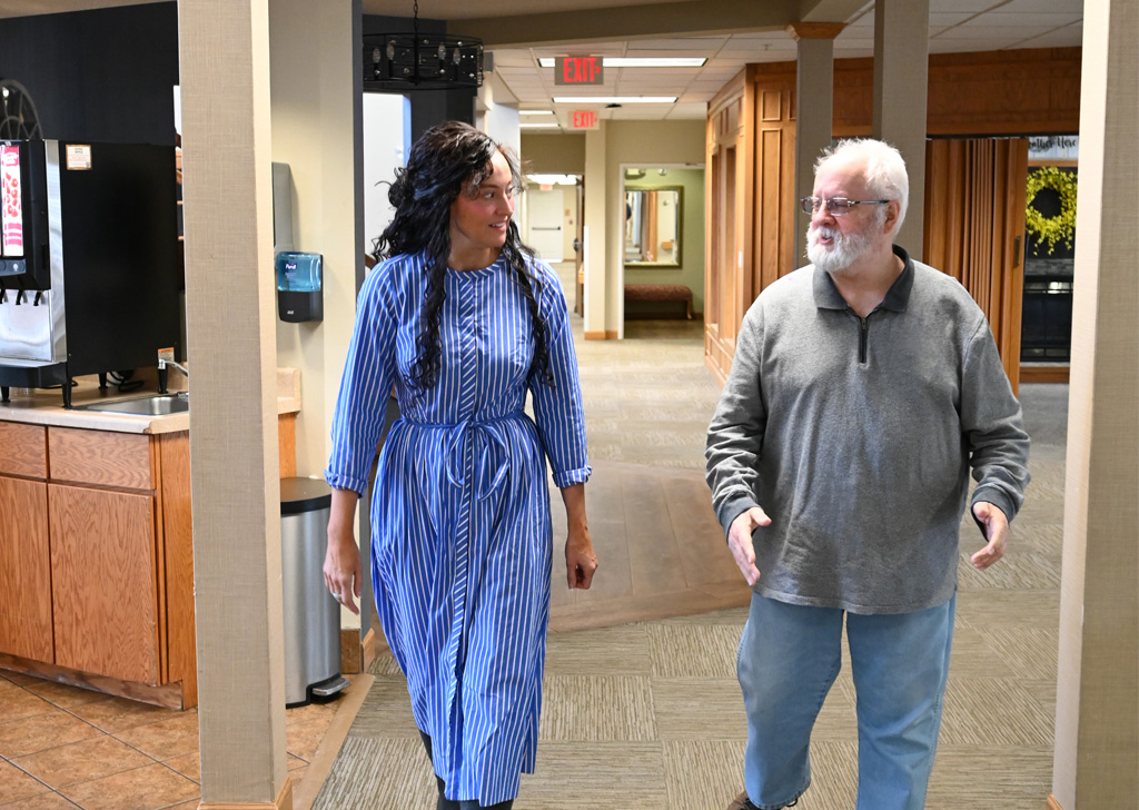 Two people walk side by side down a well-lit hallway, talking, with a coffee station and wooden cabinetry visible along the wall and exit signs overhead.